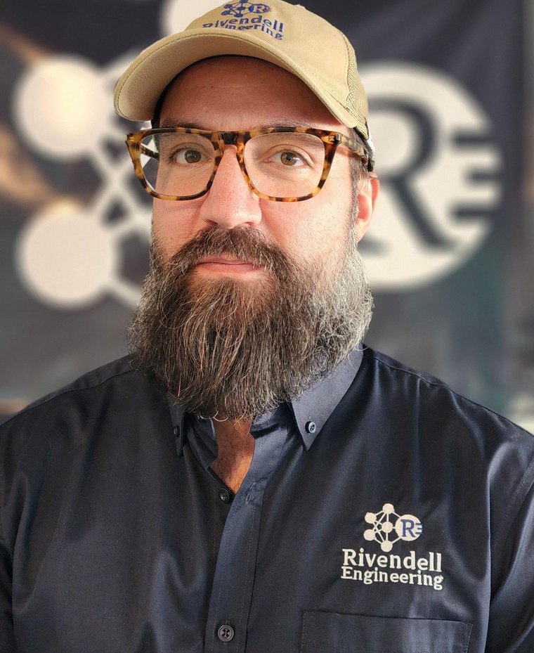 Craig Olivo, founder of Rivendell Engineering, wearing a Rivendell Engineering shirt and cap in front of a banner carrying the company mark.